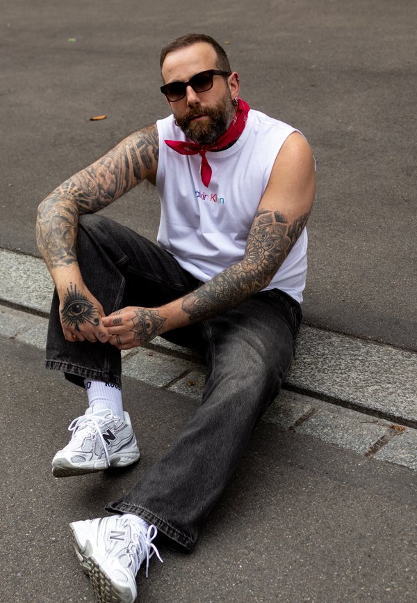 Balta be rankovių marškiniai su "Calvin Klein" logotipu, raudona bandana, juodos džinsai, baltos New Balance sportbačiai ir išsamios tatuiruotės ant rankų.