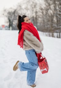 Femme portant une écharpe rouge, une veste en fourrure beige et un jean, debout sur une jambe dans la neige, tenant un sac à main rouge avec des motifs de graffiti.