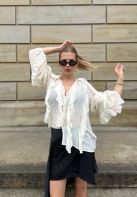 Blouse blanche à volants avec de longues manches bouffantes et des boutons, associée à une jupe noire avec une fente, devant un mur en pierre texturé.
