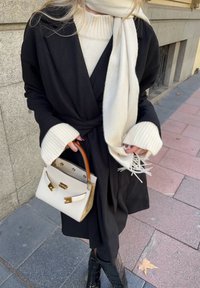 Μαύρο παλτό με φαρδιά μανίκια, κρεμ πουλόβερ με ρίγες, λευκό κασκόλ �και μια μικρή λευκή τσάντα με καφέ λαβή και χρυσές λεπτομέρειες.