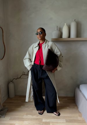 Mujer con abrigo beige tipo gabardina, camiseta roja, pantalones negros anchos, gafas de sol, sosteniendo un bolso grande tejido, de pie en una habitación minimalista con jarrones en la estantería.
