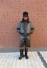 Fekete bőr bomberdzseki fekete kapucnis pulóver fölött, térdig érő bőr rövidnadrággal és fekete gumicsizmával, téglafal előtt.