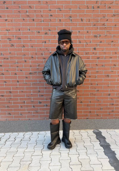 Cazadora bomber de cuero negro sobre una sudadera negra con capucha, combinada con pantalones cortos de cuero hasta la rodilla y botas de goma negras, frente a una pared de ladrillos.