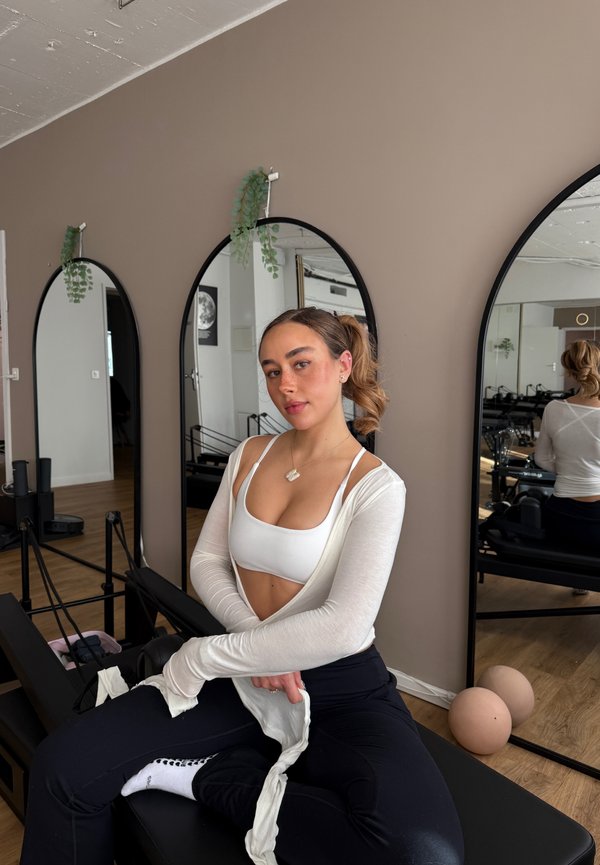 Joven mujer con ropa deportiva sentada en una máquina de Pilates reformer en un estudio con tres espejos arqueados y pelotas de ejercicio en un suelo de madera.