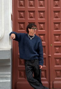 Maglione oversize blu navy con texture, pantaloni ampi in denim scuro, collana in argento spesso, occhiali da sole rotondi e auricolari wireless.