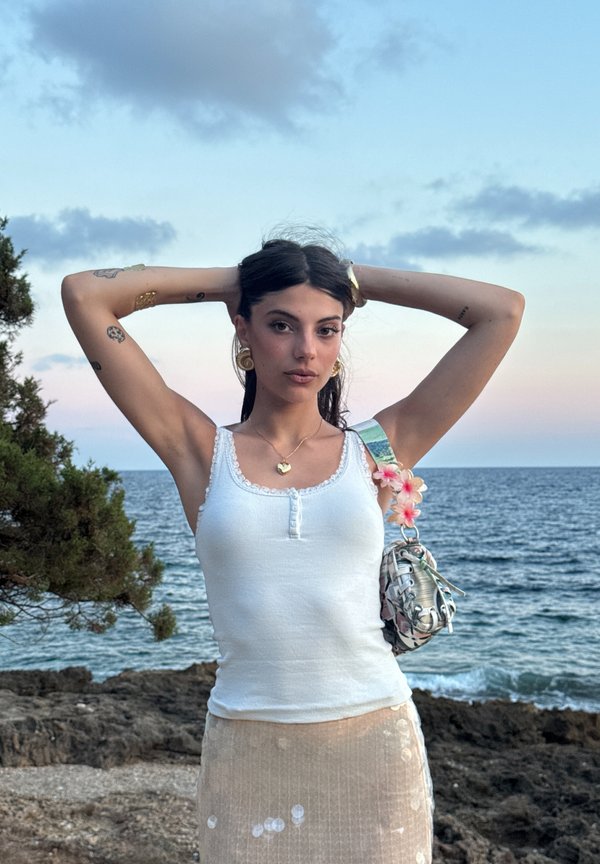 Jeune femme tatouée portant un débardeur blanc et une jupe beige, debout les mains sur la tête près d'une côte rocheuse et de la mer au coucher du soleil.