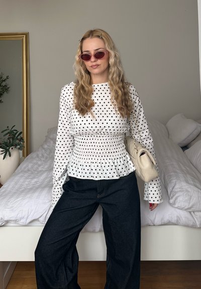 Mujer con cabello rubio ondulado que lleva gafas de sol rosas, blusa blanca de manga larga con lunares, pantalones negros y un bolso beige acolchado de hombro.