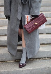 Grauer Wollmantel, burgunderfarbene gewebte Clutch, graue Socken und burgunderfarbene spitze Schuhe mit Schnallen-Detail. Stufen im Hintergrund sichtbar.