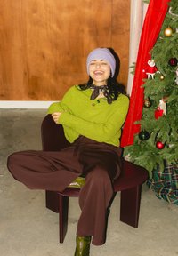 Femme souriante en pull vert, pantalon marron et bonnet violet, assise en tailleur sur une chaise à côté d'un sapin de Noël décoré avec un rideau rouge.