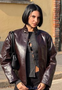 Burgundska umjetna kožna jakna s sjajnim završetkom, dizajnirana s ovratnikom i zatvaranjem na cipzar, nošena preko sivog majice s gumbima.