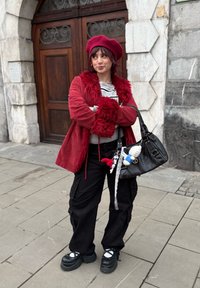 Manteau en fausse fourrure rouge sur un haut rayé, pantalon cargo noir et chaussures plateformes noires épaisses. Sac à main noir avec des breloques décoratives. Béret.