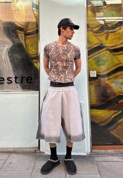 Camiseta ajustada de manga corta con estampado de camuflaje; pantalones cortos gris holgados con bolsillos; calcetas negras; zapatos negros sin cordones. Gorra negra usada al revés.