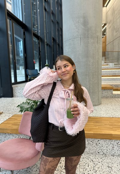 Joven mujer con blusa rosa y medias estampadas sostiene una bebida verde, parada cerca de una escalera moderna y una silla rosa en un espacio interior luminoso.