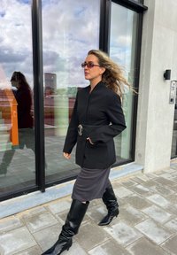Blazer nero su misura con rever ampio, vita stretta e dettaglio in argento. Abbinato a una gonna midi grigia e stivali neri sopra il ginocchio.