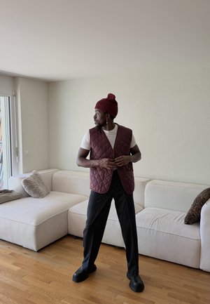 Hombre con gorro rojo, chaleco acolchado, camiseta blanca y pantalones de rayas de ganchillo, de pie en una sala de estar con sofá seccional blanco y suelo de madera.