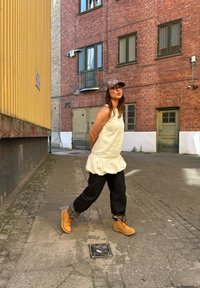 Uformell antrekk bestående av en kremfarget topp med volanger, svarte bukser og brune støvler. Bakgrunnen har røde murvegger og en gul bygning.
