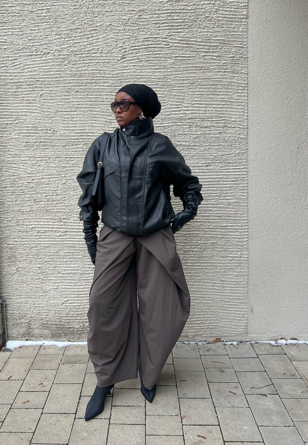Schwarze Lederjacke mit hohem Kragen, kombiniert mit lockeren, drapierten braunen Hosen. Schwarze Schuhe und übergroße Sonnenbrillen vervollständigen das Outfit.