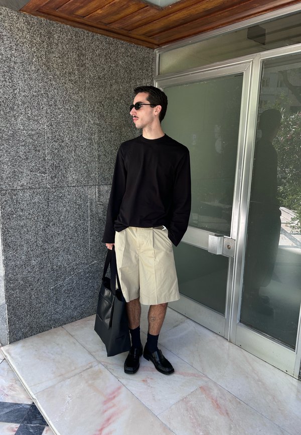Man met een zonnebril, draagt een zwart hemd met lange mouwen, beige short, zwarte schoenen, sokken, draagt een zwarte draagtas, staat bij een glazen deur.