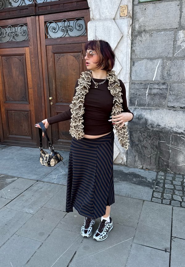Person wearing a dark brown top, beige ruffled scarf, striped skirt, trainers, sunglasses, holding a small patterned handbag, standing on a stone pavement.