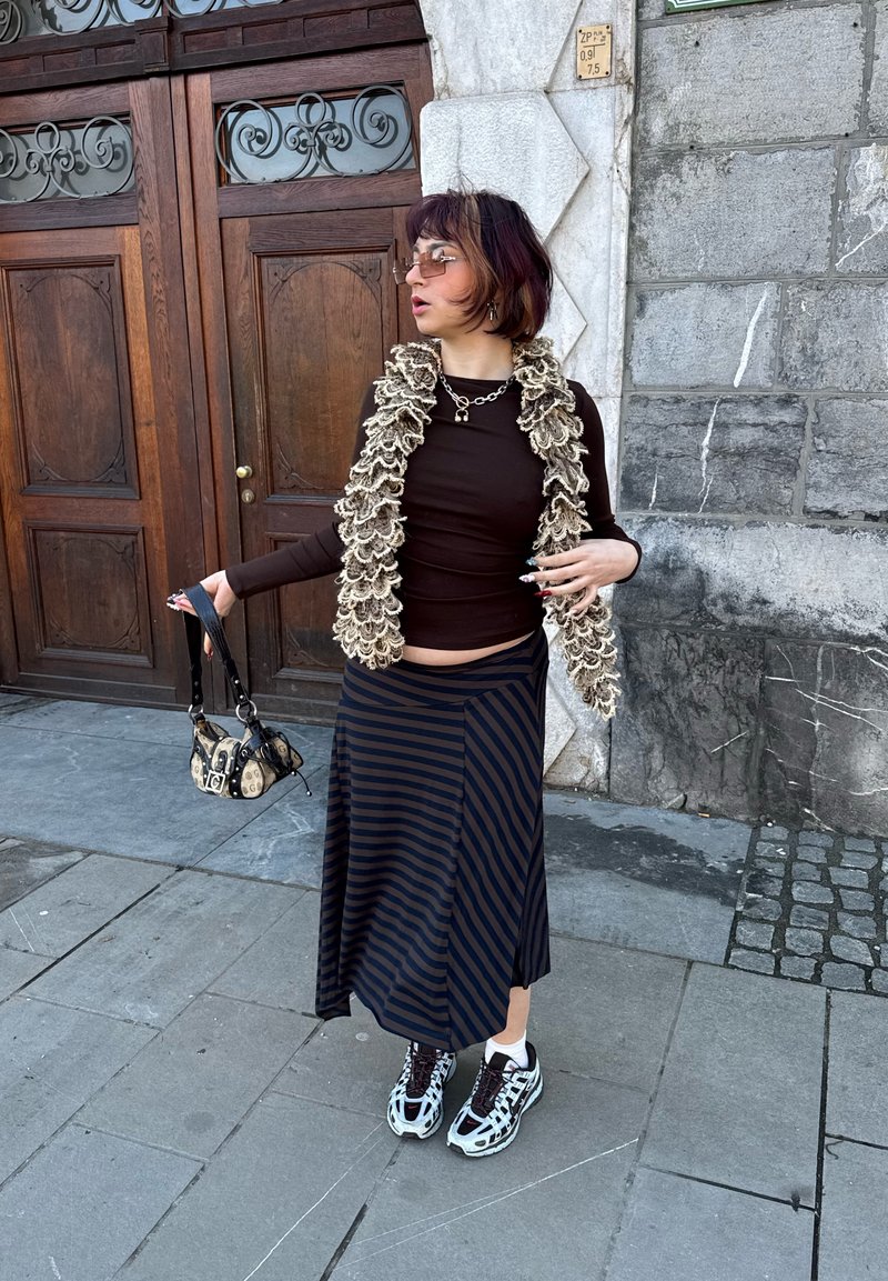 Personne portant un haut marron foncé, une écharpe beige à volants, une jupe rayée, des baskets, des lunettes de soleil, tenant un petit sac à main à motifs, debout sur un trottoir en pierre.