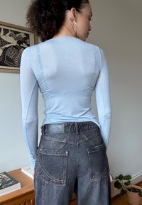 Femme aux cheveux bouclés portant un haut à manches longues bleu clair et un jean en denim foncé taille haute, debout à l'intérieur près d'un meuble en bois et d'une plante.