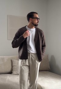 Homme portant des lunettes, un cardigan marron, une chemise blanche et un pantalon crème, debout près d'un canapé beige dans une pièce minimaliste.