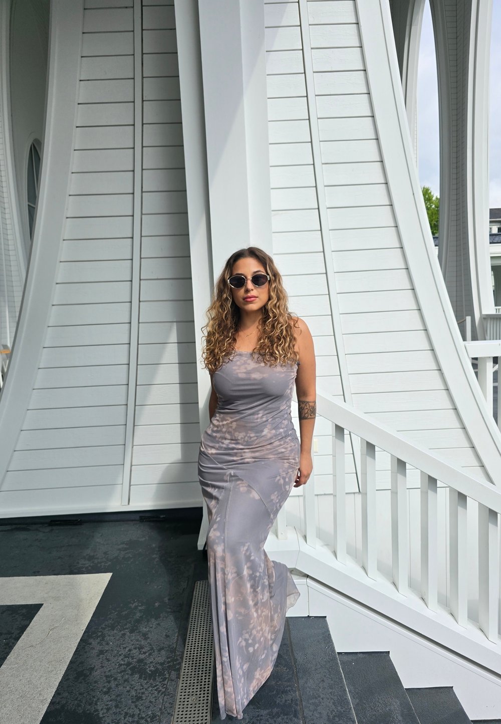 Dlhé priliehavé sivé šaty s jemným batikovým vzorom. Kučeravé vlasy a okrúhle slnečné okuliare. Stojí na schodoch blízko bielych architektonických prvkov.