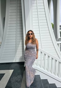 Dlhé priliehavé sivé šaty s jemným batikovým vzorom. Kučeravé vlasy a okrúhle slnečné okuliare. Stojí na schodoch blízko bielych architektonických prvkov.