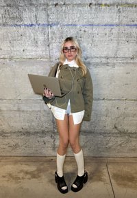 Blond kvinne iført solbriller, grønn jakke, hvite sokker og platåsko holder en åpen laptop og står foran en betongvegg.