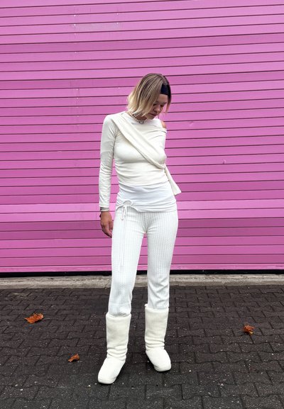 Top blanco de manga larga con diseño drapeado, pantalones blancos texturizados y botas blancas esponjosas, frente a una pared de madera rosa.