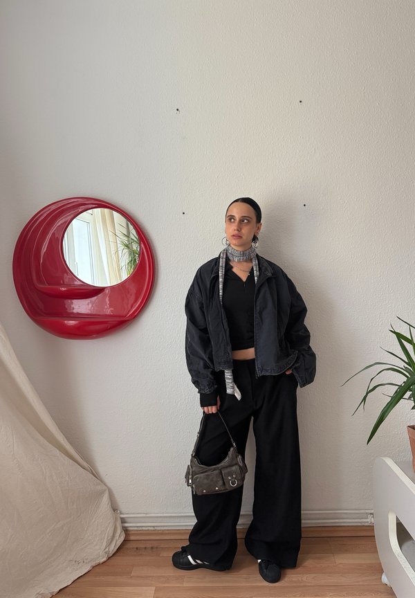 Čierna skrátená bomber bunda cez čierny top v kombinácii s voľnými čiernymi nohavicami. Drží malú šedú kabelku. Na stene červené kruhové zrkadlo.