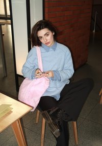 Suéter oversized azul claro com uma camisa xadrez de colarinho por baixo, calças pretas e uma bolsa rosa fofa; usando botas pretas, sentado em um café.