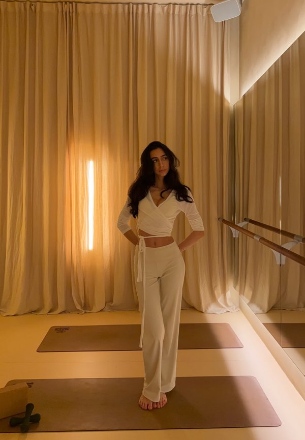 Kvinde i beige bukser og omslags-top står barfodet på yogamåtte i blødt oplyst rum med gardiner fra gulv til loft og vægspejl med balletstænger.