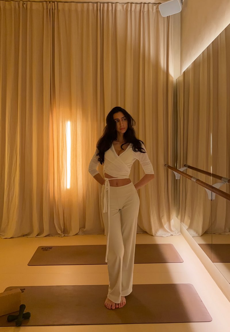 Femme en pantalon beige et haut cache-cœur, debout pieds nus sur un tapis de yoga dans une pièce doucement éclairée avec des rideaux du sol au plafond et un miroir mural avec des barres de ballet.