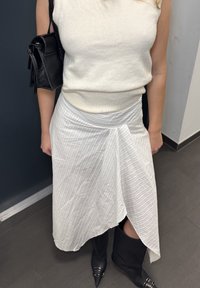 Jupe portefeuille blanche à texture à rayures verticales, associée à un pull crème ajusté et des bottes noires montant jusqu'au genou. Petit sac à main noir en accent.