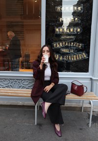 Blazer color mogano, top bianco e pantaloni neri a gamba larga indossati da una figura seduta. Tiene in mano una tazza di caffè, con una borsa bordeaux accanto.
