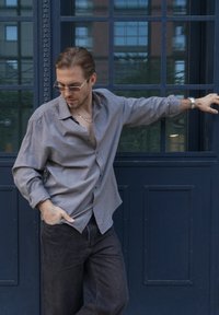 Man met een bril, een grijze shirt en donkere broek leunt tegen een donkerblauwe deur, met één hand in zijn zak en de andere uitgestrekt.