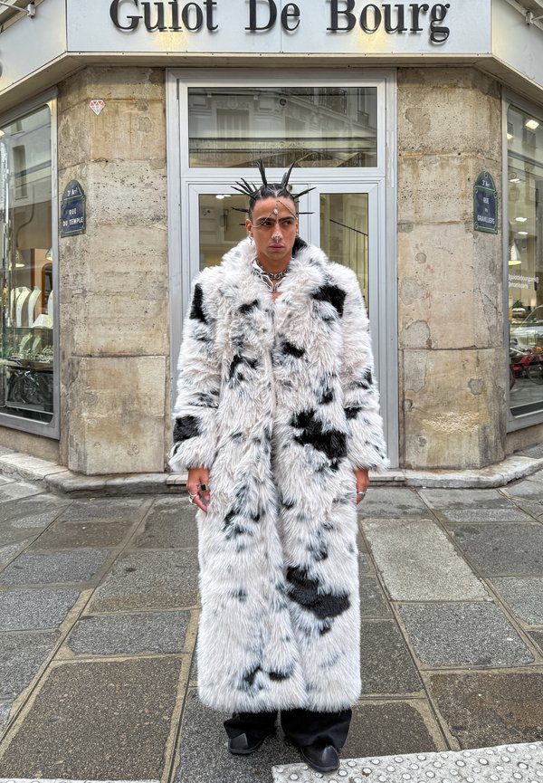 Person with spiked hair and facial jewelry wearing a long white and black fur coat stands in front of a stone building with glass doors.