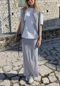 Grijze mouwloze cardigan over een witte t-shirt, gecombineerd met een grijze maxi rok en witte sneakers met grijze accenten. Gelaagde stenen achtergrond.