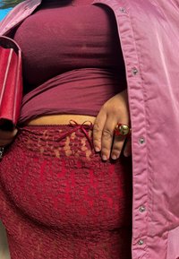 Gonna rossa in pizzo con dettaglio a fiocco, abbinata a un top bordeaux e a una giacca rosa. Un anello rosso lucido è indossato sulla mano che tiene una borsa a sorpresa coordinata.