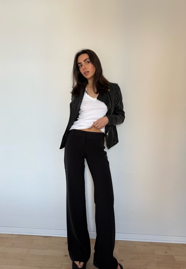 Femme aux longs cheveux foncés portant une veste en cuir noire, un t-shirt blanc et un pantalon noir à jambes larges, debout contre un mur clair uni.