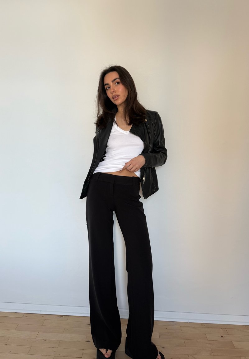 Femme aux longs cheveux foncés portant une veste en cuir noire, un t-shirt blanc et un pantalon noir à jambes larges, debout contre un mur clair uni.