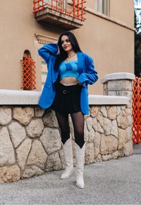 Blazer oversize bleu, top court en tricot turquoise, jupe plissée noire, collants transparents et bottes de cowboy blanches. Fond en mur de pierre.