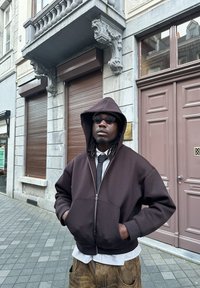 Brauner Reißverschluss-Hoodie mit Kapuze, über einem weiß-blau gestreiften Hemd und einer Krawatte getragen, kombiniert mit beigen Hosen. Strukturiertes Material, lässiger Schnitt.