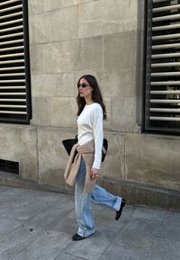 Biała bluzka z długim rękawem, jasnoniebieskie rozszerzane dżinsy, beżowy dziergany pas w talii, czarna torba, czarne buty, okulary przeciwsłoneczne. Spacer przy kamiennym murze.