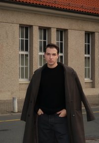 Casaco escuro com grandes lapelas sobre um suéter preto, combinado com jeans escuros. O fundo apresenta grandes janelas e paredes de pedra texturizada.