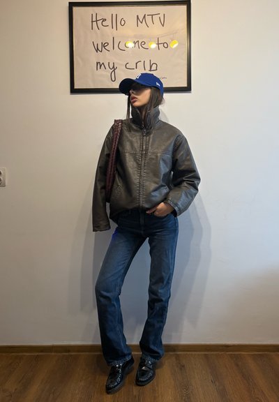 Joven con gorra azul, chaqueta de cuero marrón y vaqueros, de pie junto a una pared con un cartel enmarcado que dice "Hola MTV, bienvenidos a mi hogar."