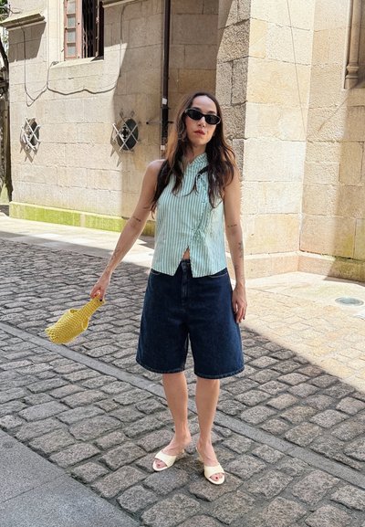 Top sin mangas de rayas verde claro con detalle de nudo, pantalones cortos de mezclilla oscuros, bolso tejido amarillo y sandalias deslizables blancas en una calle de adoquines.