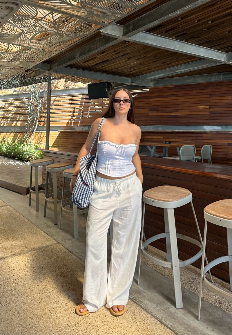 Top bianco senza spalline con dettaglio annodato, pantaloni lunghi in lino bianco svolazzanti e borsa a scacchi blu. Sullo sfondo si vedono pannelli di legno e sgabelli da bar.