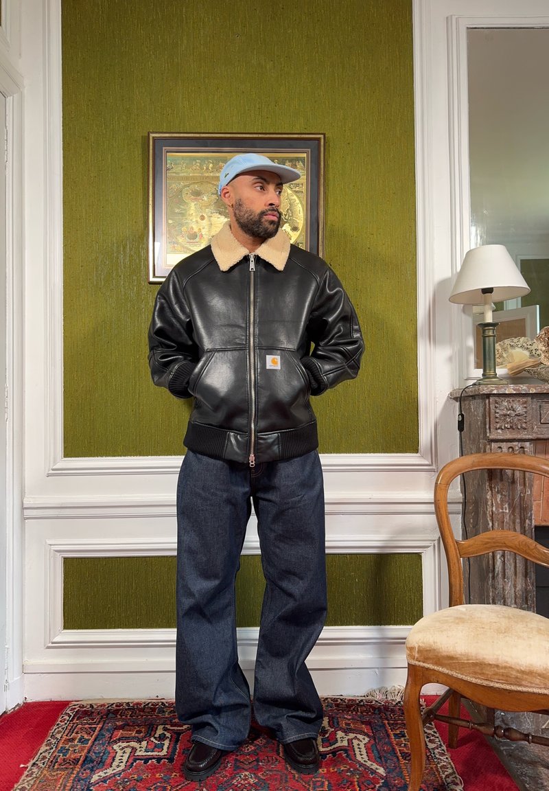 Mand iført lyseblå kasket, sort læderjakke med fleecekrave, vide jeans, stående i et rum med grøn væg og vintage møbler.